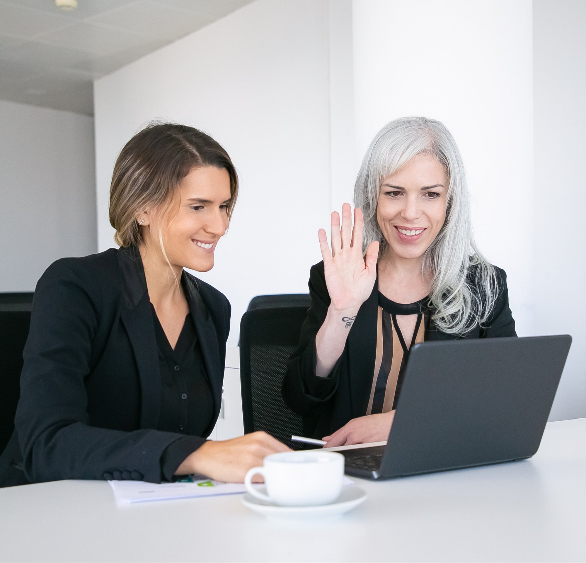 two-cheerful-female-colleagues-using-laptop-video-call-sitting-table-with-cup-coffee-looking-display-waving-hello-online-communication-concept two-cheerful-female-colleagues-using-laptop-video-call-sitting-table-with-cup-coffee-looking-display-waving-hello-online-communication-concept